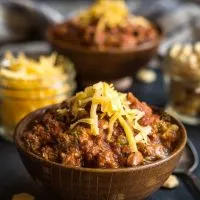 A brown bowl heaped with beef chili and garnished with cheddar cheese sits in front of a jar of cheese, crackers, and a second bowl of chili.