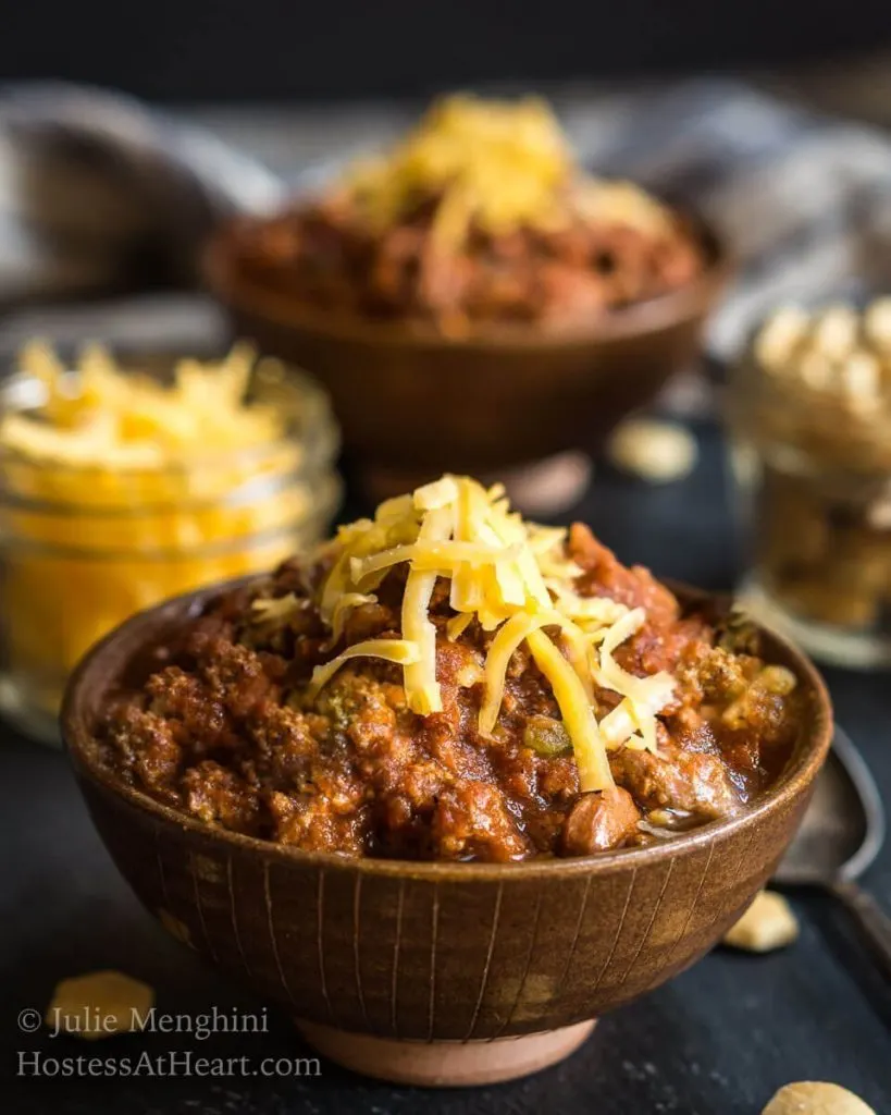 A center brown bowl heaped with chili that\'s garnished with cheddar cheese. The chili sits in front of the second bowl of chili and jars of cheese and soup crackers.