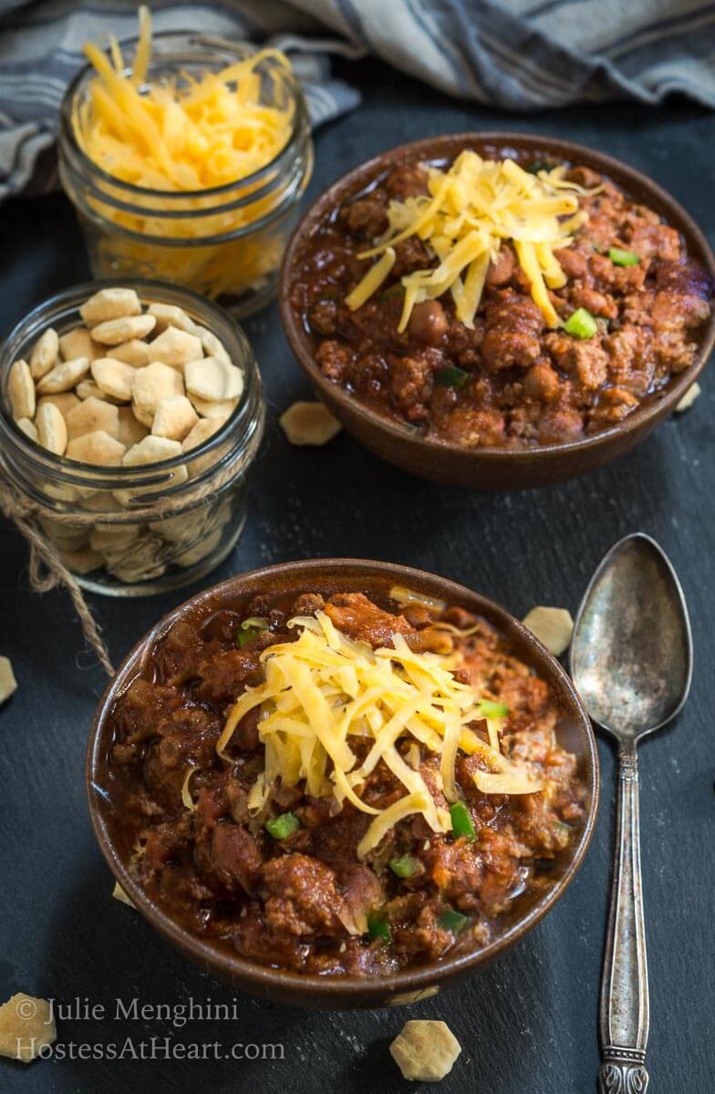 Top-down photo of two bowls of chili topped with shredded cheese in brown bowls next to small jars of cheese and soup crackers, and a spoon sitting on a slate tile.
