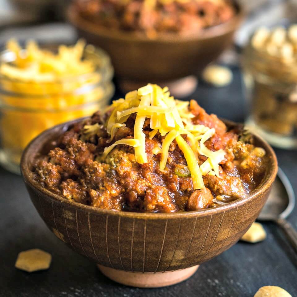 Side view of a brown bowl heaped with beef chili that\'s topped with cheddar cheese. Jars of cheese and soup crackers and a second bowl of chili sit in the background.