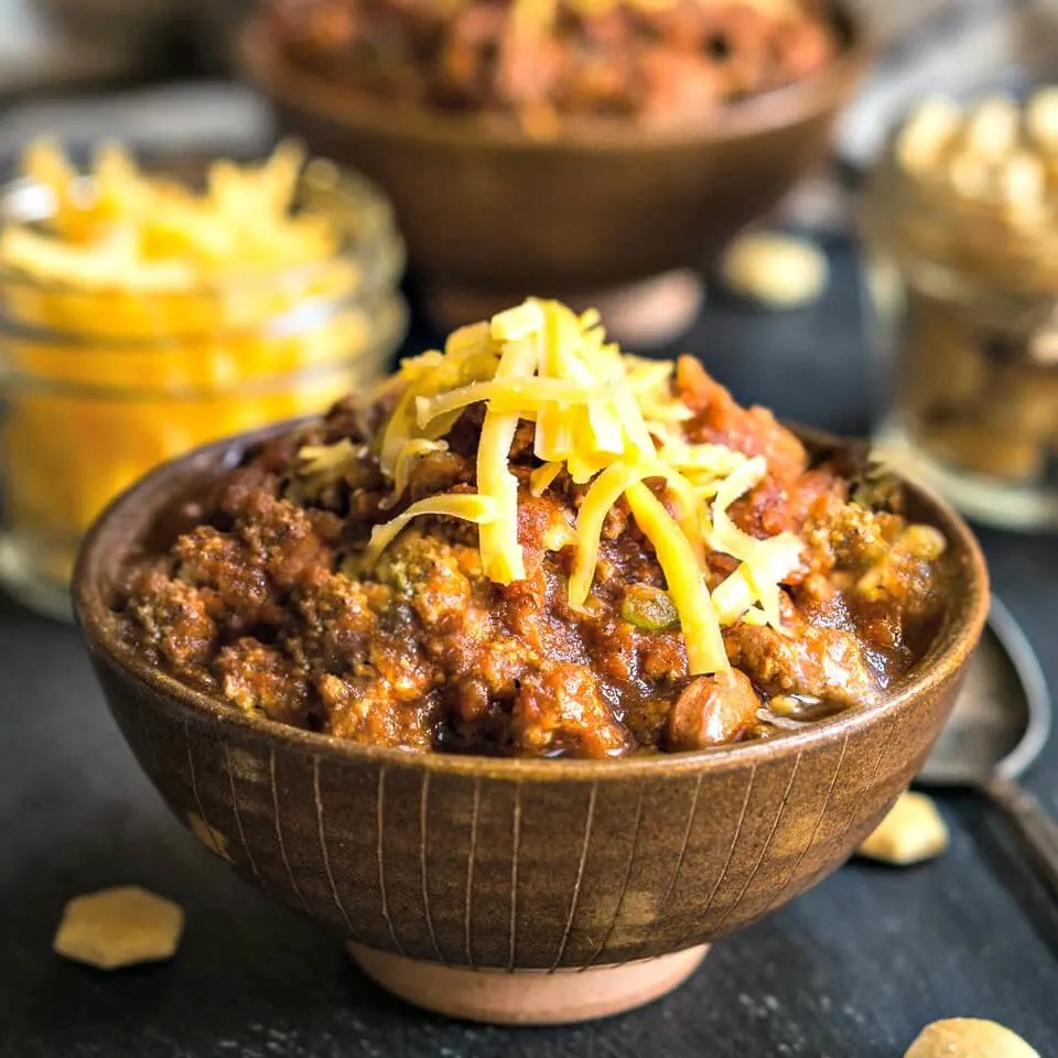 Side view of a brown bowl heaped with beef chili that\'s topped with cheddar cheese. Jars of cheese and soup crackers and a second bowl of chili sit in the background.