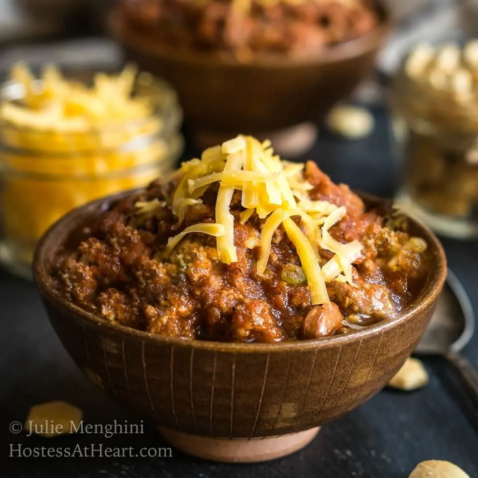 Side view of a brown bowl heaped with chili. Shredded cheese garnish the chili. Crackers and more cheese sit in jars in the background.
