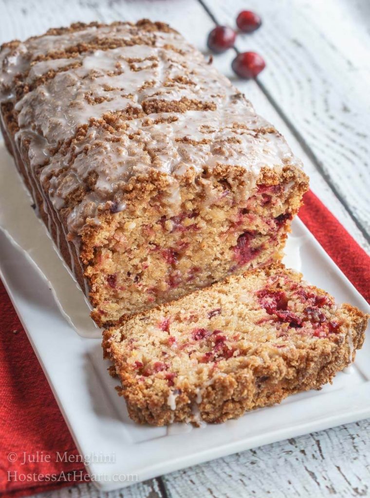 Angled view of 3/4 a loaf of cranberry apple streusel bread on a white plate over a red napkin. A slice sits in front showing the baked cranberries running through the bread.