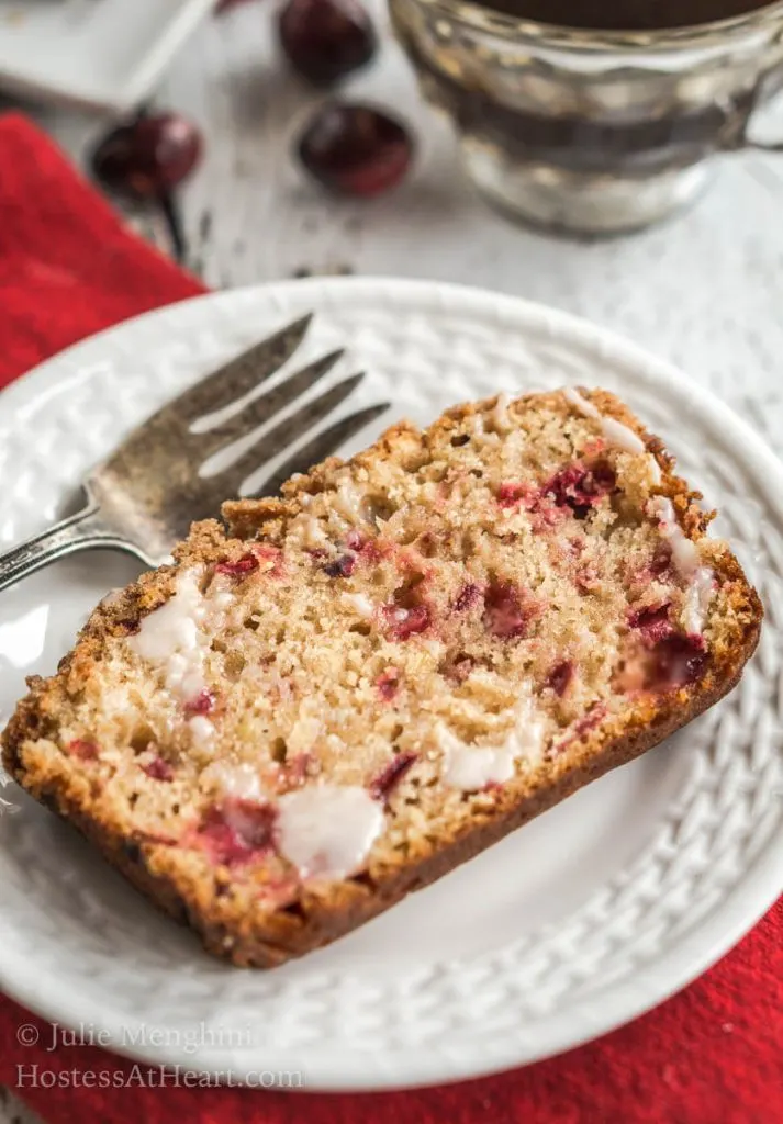 A slice of Cranberry Apple bread slathered with melting butter sitting on a white plate next to a fork over a red napking
