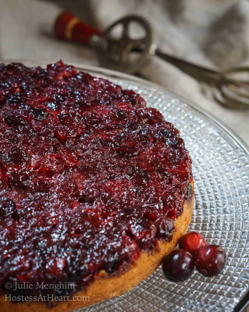 A top-down photo of a cranberry upside-down cake sitting on a glass plate. Three fresh cranberries sit next to the cake.