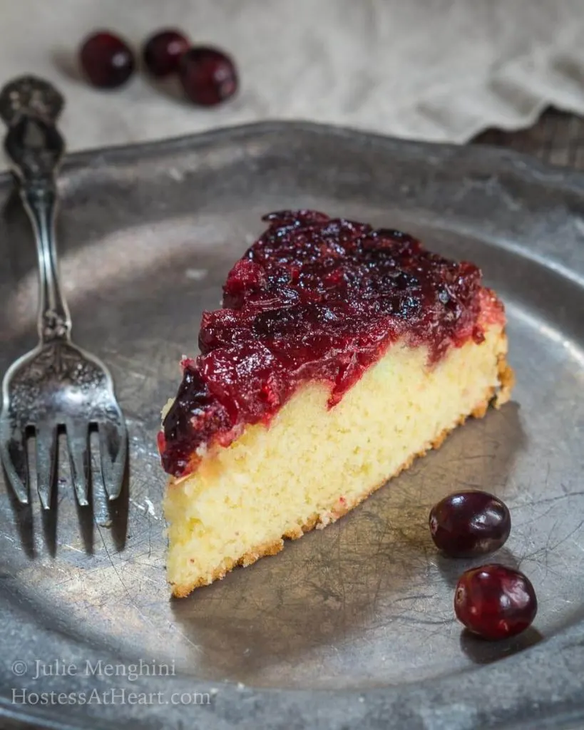 Top angle photo of a piece of soft yellow cake that\'s topped in a fresh cranberry sauce on a metal plate. 3 fresh cranberries and a fork sit on the plate next to the cake.