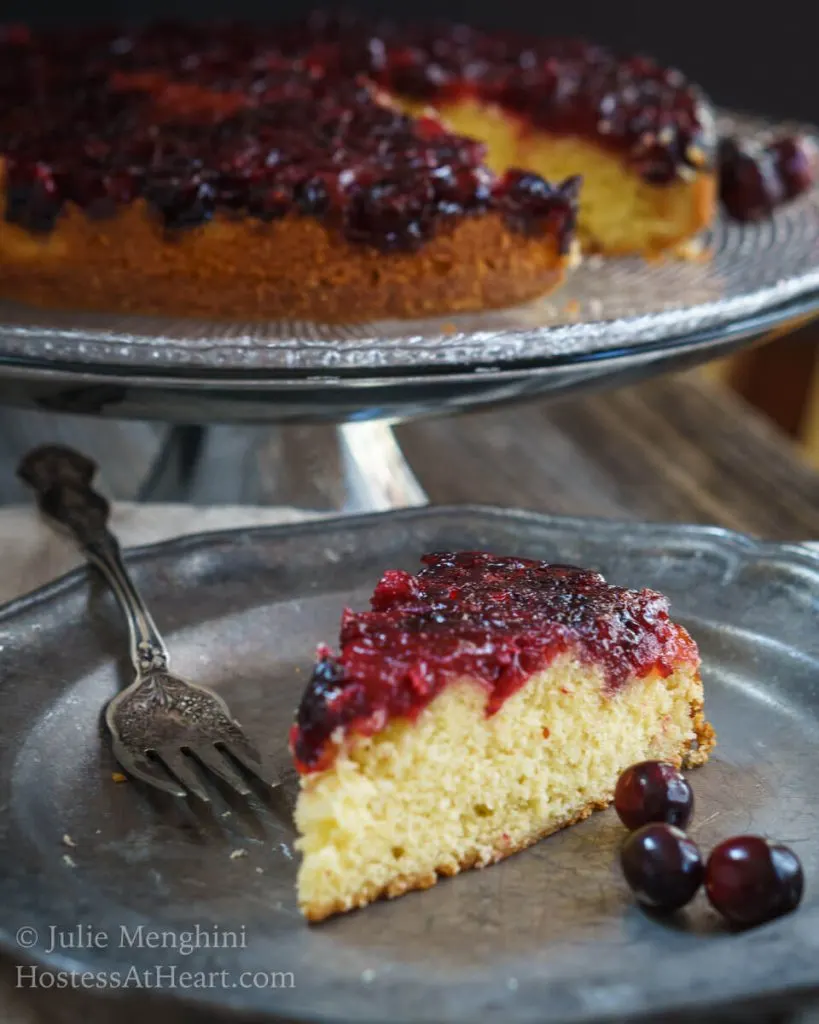 Top angle photo of a piece of soft yellow cake that\'s topped in a fresh cranberry sauce on a metal plate. 3 fresh cranberries sit on the plate next to the cake. A cake stand holding the whole cake sits in the background.