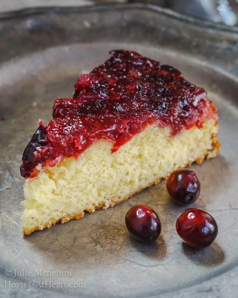 Top angle photo of a piece of soft yellow cake that\'s topped in a fresh cranberry sauce on a metal plate. 3 fresh cranberries sit on the plate next to the cake.