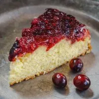 Top angle photo of a piece of soft yellow cake that's topped in a fresh cranberry sauce on a metal plate. 3 fresh cranberries sit on the plate next to the cake.