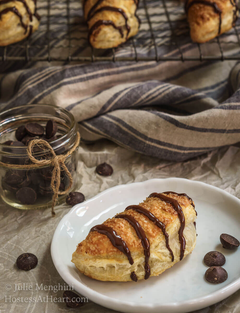 A white plate holds a puff pastry crescent that\'s been filled and drizzled with chocolate. A jar of a small jar of chocolate chips sits in the background in front of a striped blue towel.