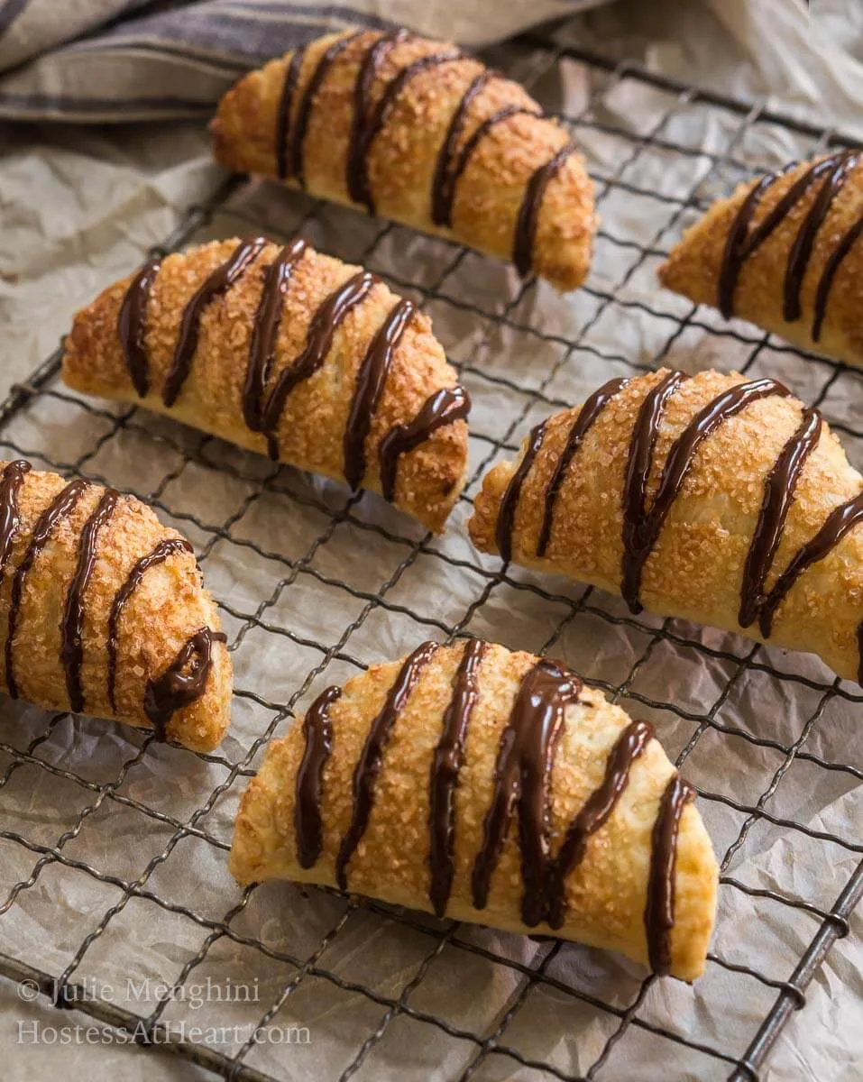 6 puff pastry crescents filled and drizzled with chocolate sit on a cooling rack over a piece of parchment paper.
