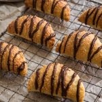 6 puff pastry crescents that are filled and drizzled with chocolate sit on a cooling rack over a piece of parchment paper.