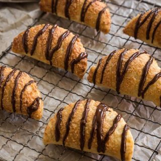 6 puff pastry crescents that are filled and drizzled with chocolate sit on a cooling rack over a piece of parchment paper.
