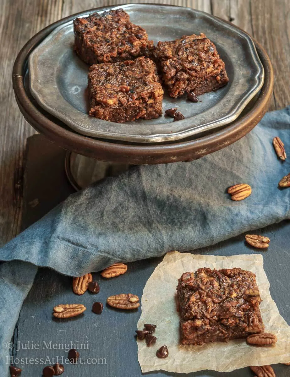 A top-down photo of a Derby Pie Brownie Bar sitting on a piece of parchment paper over a piece of slate. A cake stand holds additional bars in the background over a blue napkin. Fresh pecans and chocolate chips surround the bar.