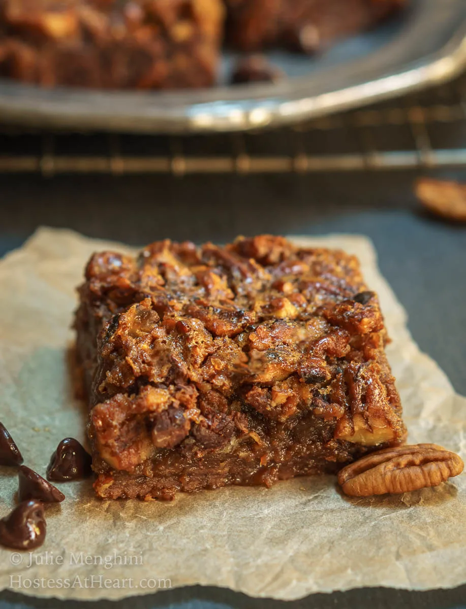 A side photo of a Derby Pie Brownie Bar sitting on a piece of parchment paper over a piece of slate. A cooling rack holds additional bars in the background.