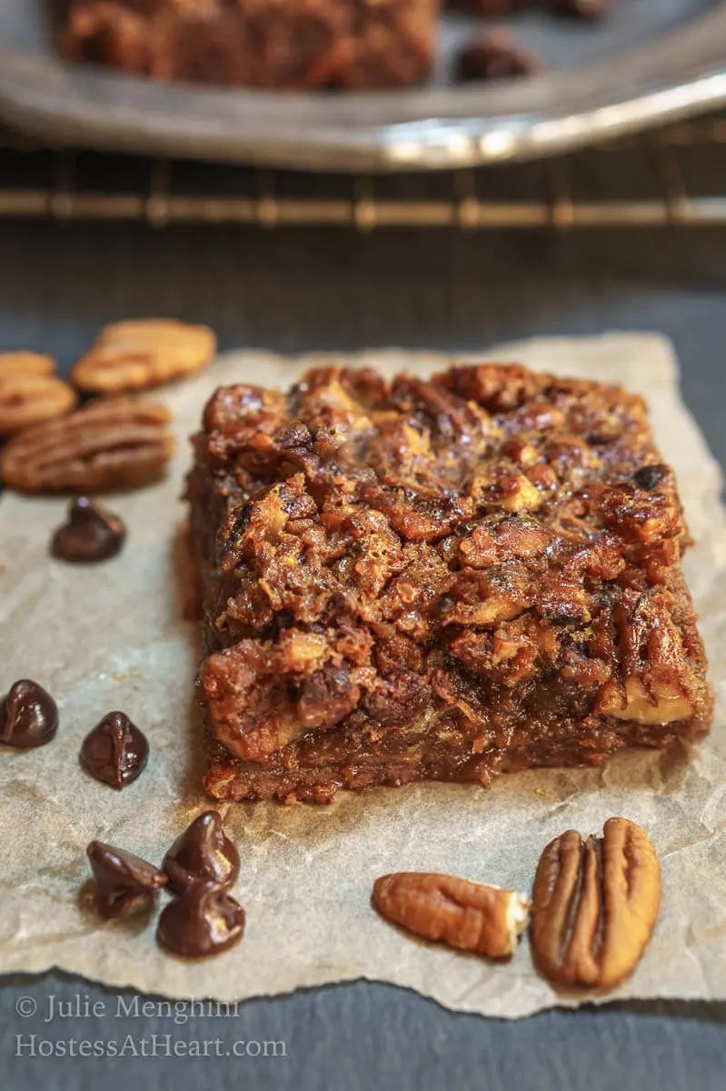 A side photo of a Derby Pie Brownie Bar sitting on a piece of parchment paper over a piece of slate. Pecans and chocolate chips sit next to the bar.