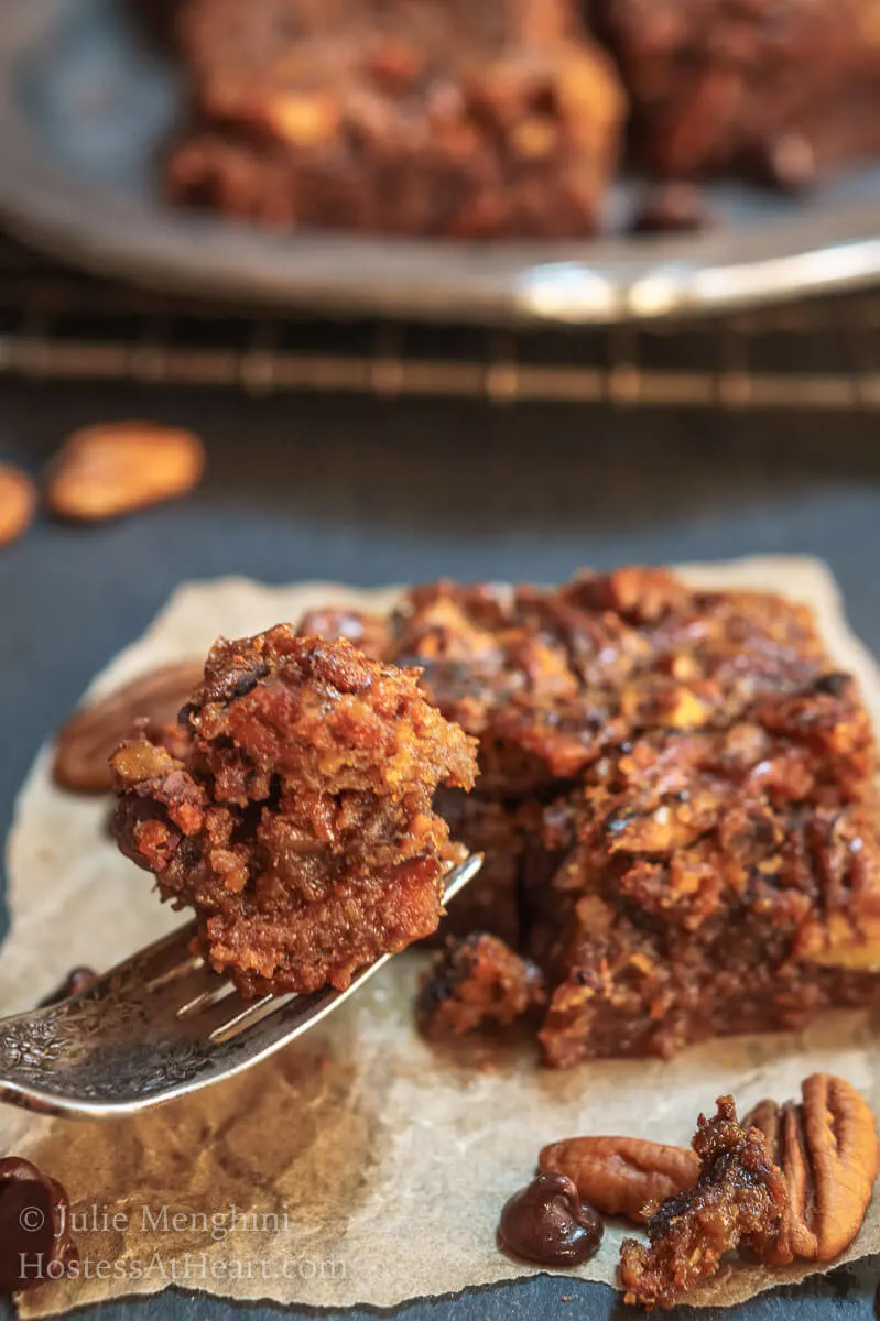 A side photo of a fork full of a Derby Pie Brownie Bar hovers over the Derby Pie Brownie that sits on a piece of parchment paper over a piece of slate. A cooling rack holds additional bars in the background.