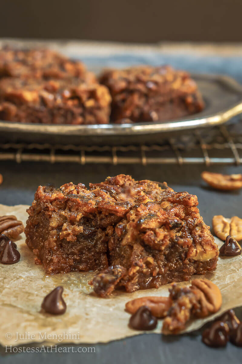 A piece of a Derby Pie Brownie sitting on a piece of parchment paper. A plate of bars sits in the background over a cooling rack.