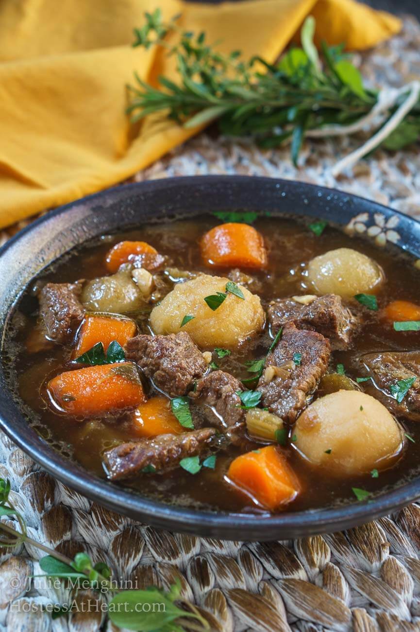 A side angle view of a bowl of stew filled with big chunks of beef and vegetables in a gray bowl over a woven placemat. A gold napkin and a bouquet of fresh herbs sit off to the side.