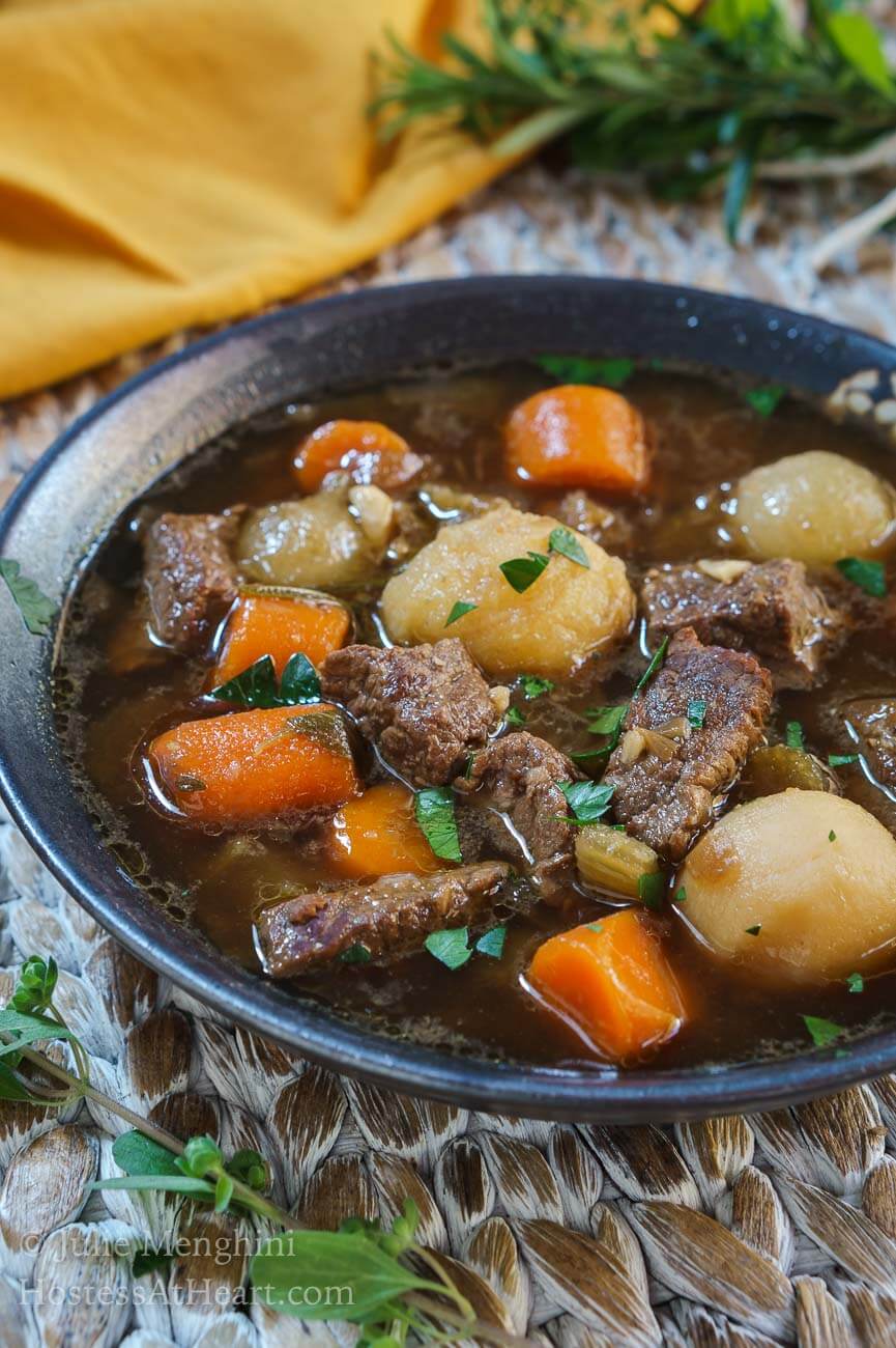 A side angle view of a partial view of a bowl of stew filled with big chunks of beef and vegetables in a gray bowl over a woven placemat. A gold napkin and a bouquet of fresh herbs sit off to the side.