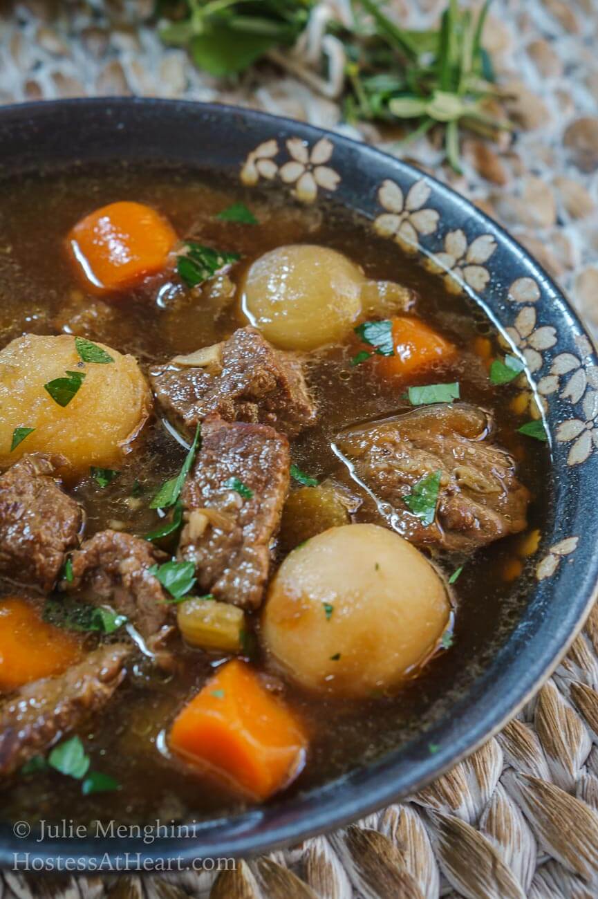 A side angle view of a partial view of a bowl of stew filled with big chunks of beef and vegetables in a gray bowl over a woven placemat. A gold napkin and a bouquet of fresh herbs sit off to the side.