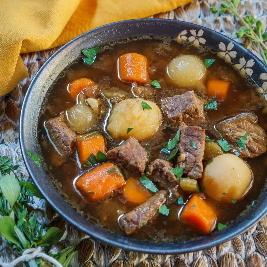 A bowl of food Vegetable Beef Soup full of carrots, potatoes, and chunks of beef in a dark gray bowl over a wooden placemat. Fresh herbs sit next to the bowl and a gold napkin sits in the background.