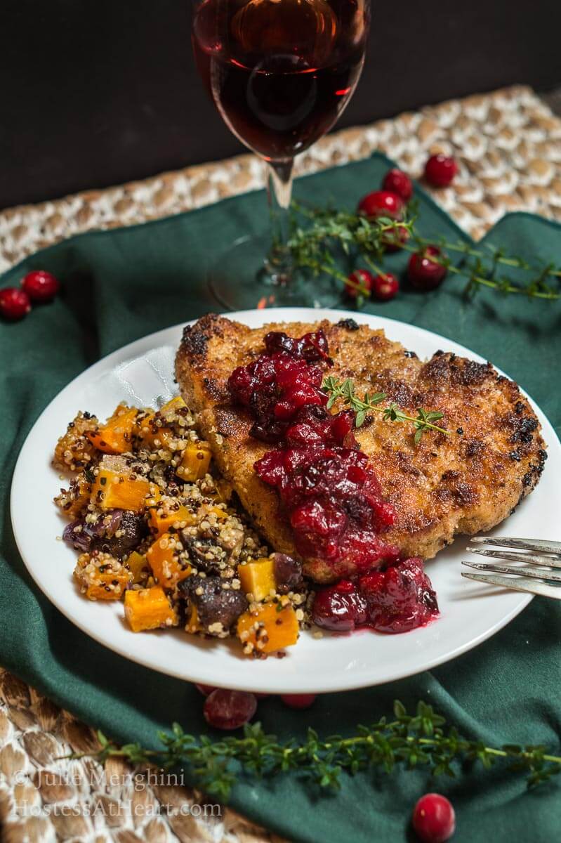 A white plate filled with a breaded Pork chop topped with a dollop of cranberry sauce and fresh thyme. Baked squash and quinoa sit next to the pork chop. The plate sits on a green napkin over a woven placemat.