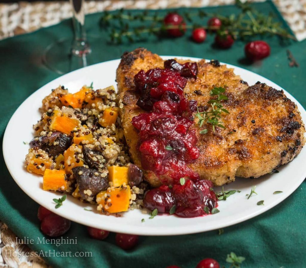 A white plate filled with a breaded Pork chop topped with a dollop of cranberry sauce and fresh thyme. Baked squash and quinoa sit next to the pork chop. The plate sits on a green napkin over a woven placemat. Fresh thyme and cranberries sit in the background.