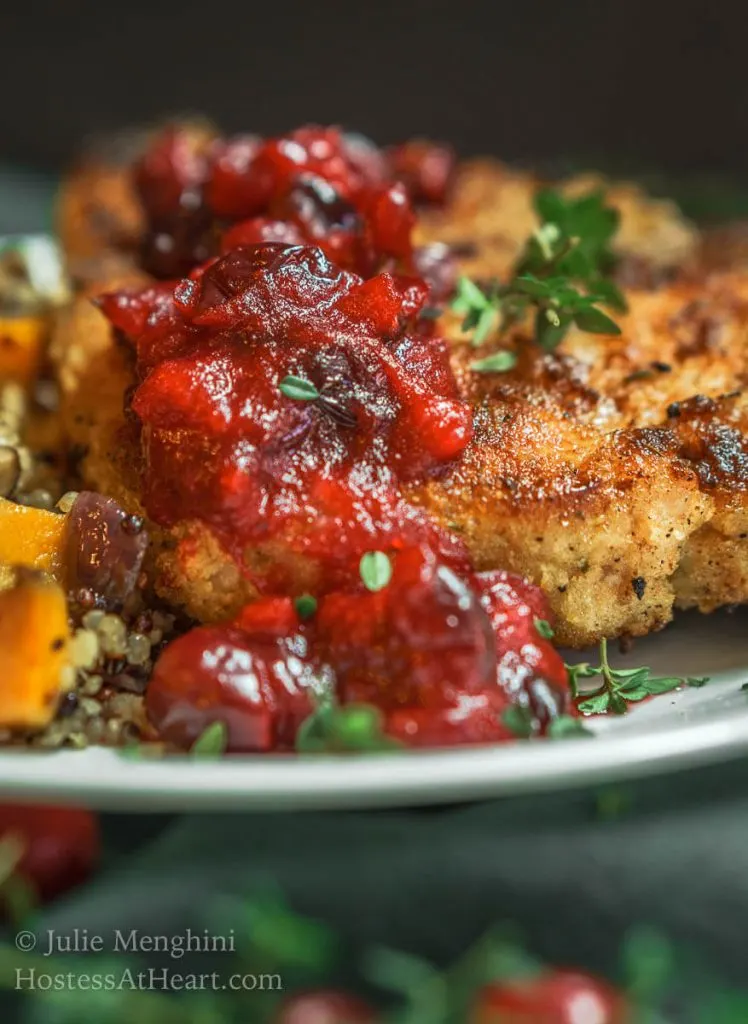 Close-up side view of a white plate filled with a breaded Pork chop topped with a dollop of cranberry sauce and fresh thyme. Baked squash and quinoa sit next to the pork chop. The plate sits on a green napkin over a woven placemat.