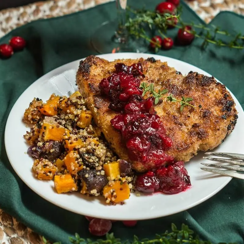 A white plate filled with a breaded Pork chop topped with a dollop of cranberry sauce and fresh thyme. Baked squash and quinoa sit next to the pork chop. The plate sits on a green napkin over a woven placemat.