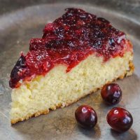 Top angle photo of a piece of soft yellow cake that's topped in a fresh cranberry sauce on a metal plate. 3 fresh cranberries sit on the plate next to the cake.