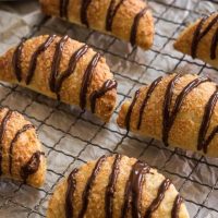 6 puff pastry crescents that are filled and drizzled with chocolate sit on a cooling rack over a piece of parchment paper.