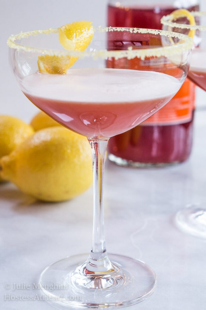 A coupe glass of an Italian cocktail called Arancia Orange Italiano. A lemon twist sits in the glass and the glass sits next to a lemon and the martini mixer is in the background.