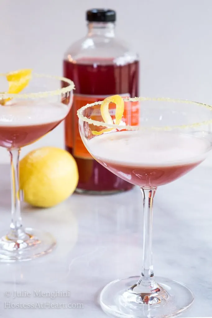 A coupe glass of an Italian cocktail called Arancia Orange Italiano. A lemon twist sits in the glass and the glass sits next to a lemon and the martini mixer is in the background.