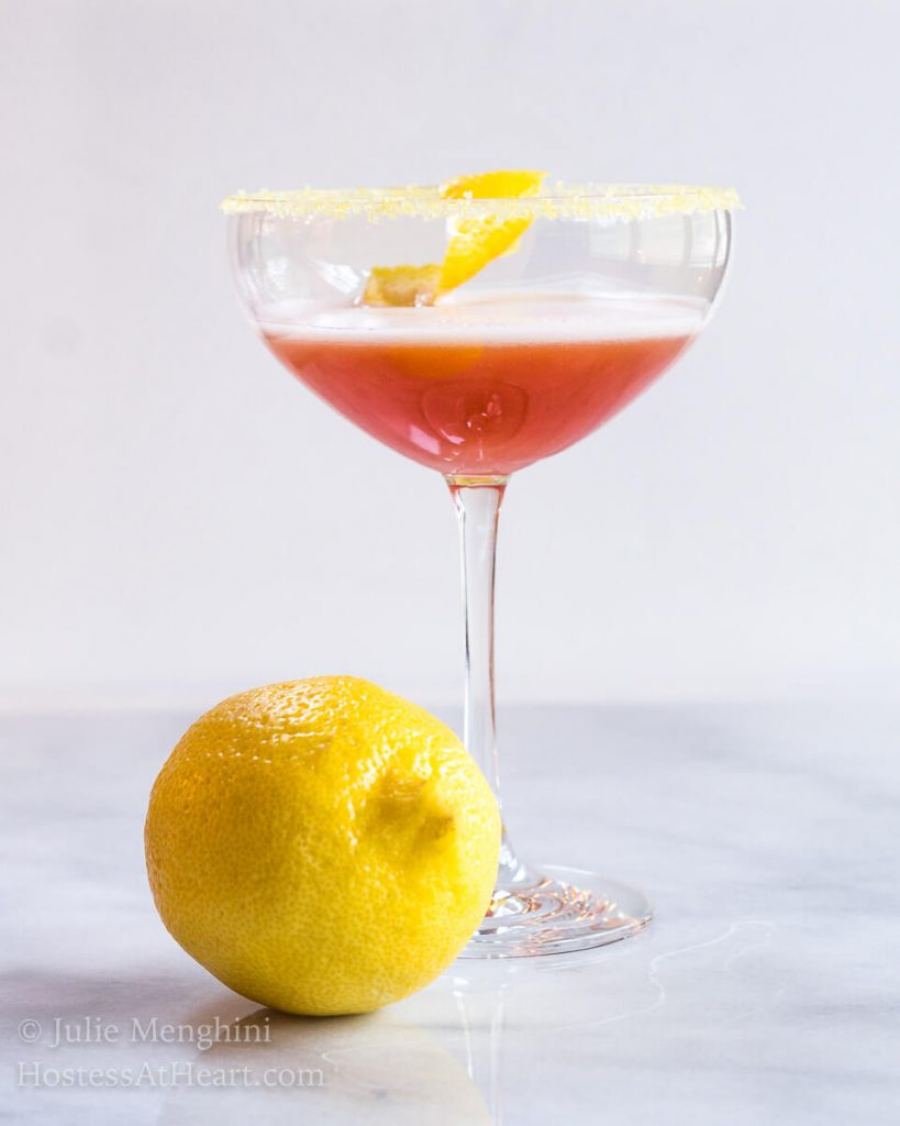 A coupe glass of an Italian cocktail called Arancia Orange Italiano. A lemon twist sits in the glass and the glass sits next to a lemon.