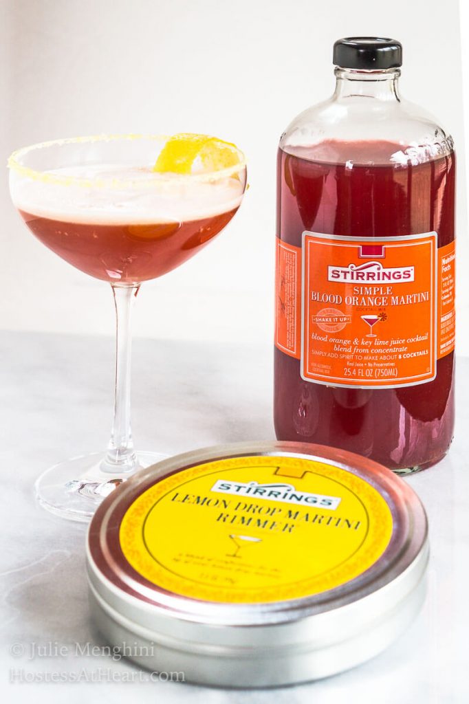 A coupe glass of an Italian cocktail called Arancia Orange Italiano. A lemon twist sits in the glass and the glass sits next to a Lemon Martini rimmer sugar and Blood Orange Martini mixer.