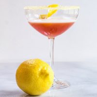 A glass Arancia Orange Italiano cocktail with fresh lemon in front of it.