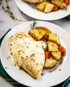 A chicken breast with a lemon herb sauce sitting on a white plate next to roasted potatoes. The plate sits over a green napkin with herbs in the background.