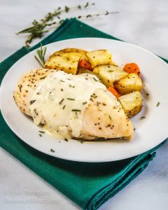 A chicken breast with a lemon herb sauce sitting on a white plate next to roasted potatoes. The plate sits over a green napkin with herbs in the background.