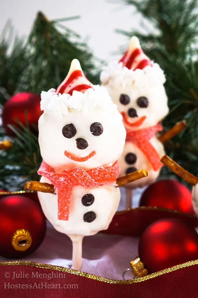 2 Rice Krispie Snowman Pops with cute little chocolate chip eyes, striped candy hat and a candy leather mufflers sit over a red ribbon with pine trees in the background.