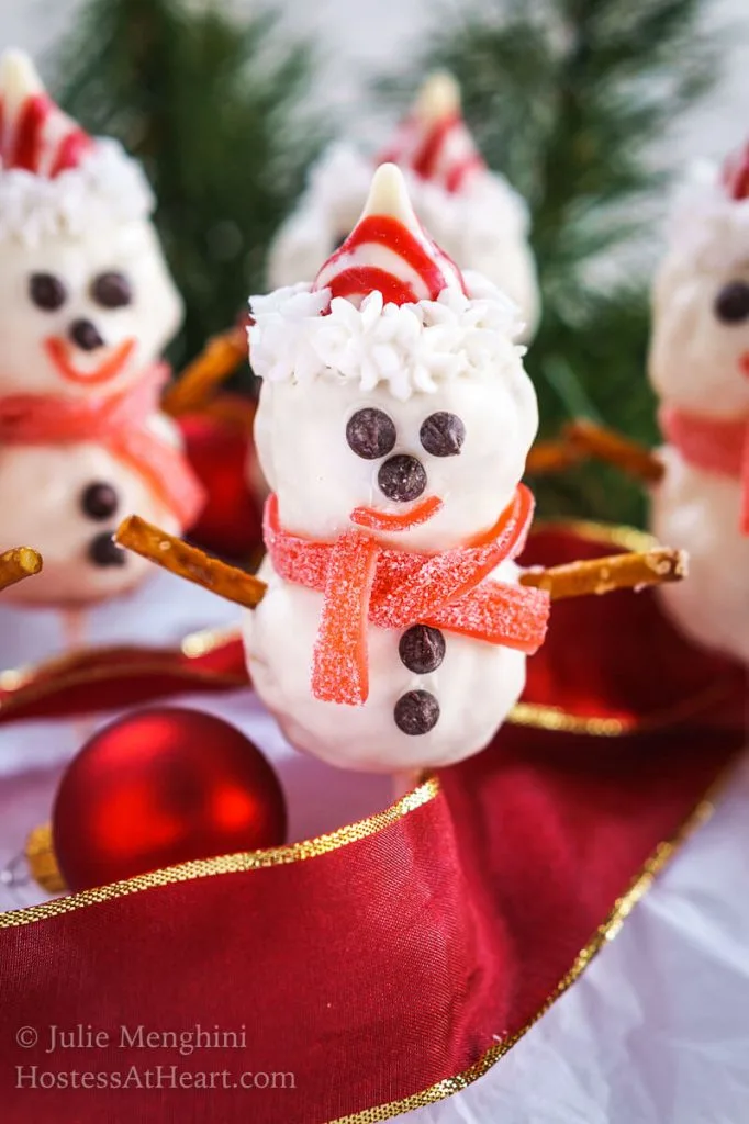 4 Rice Krispie Snowman Pops with cute little chocolate chip eyes, striped candy hat and a candy leather mufflers sit over a red ribbon with pine trees in the background.