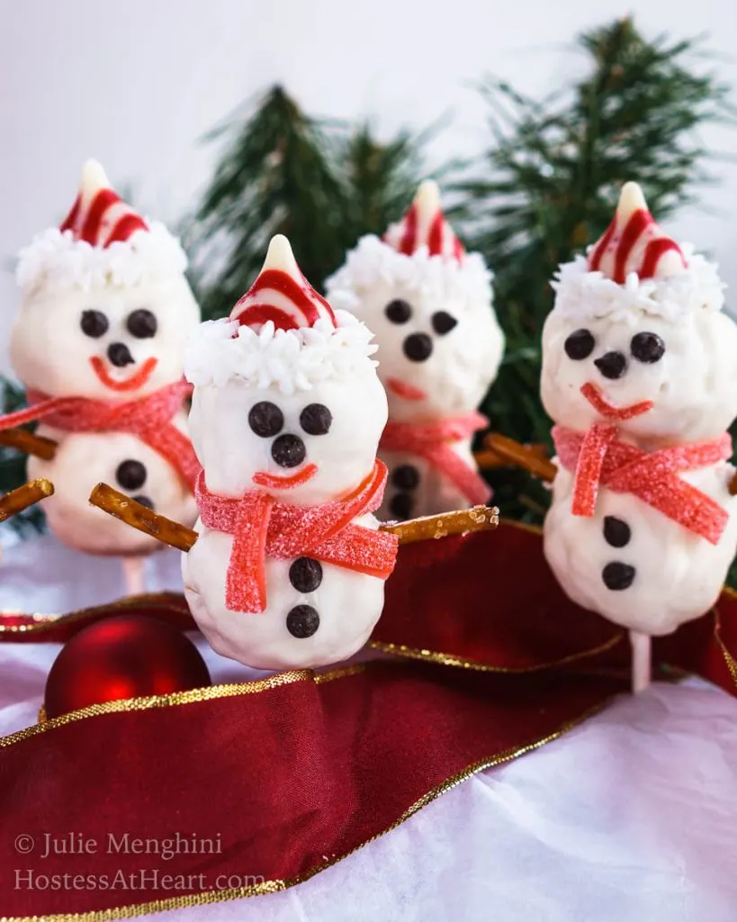 4 Rice Krispie Snowman Pops with cute little chocolate chip eyes, striped candy hat and a candy leather mufflers sit over a red ribbon with pine trees in the background.
