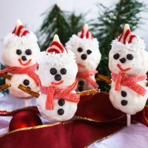 Four snowman pops with chocolate chip eyes and buttons dressed with a candied hat and muffler sitting in fron of pine trees and on top of ribbon