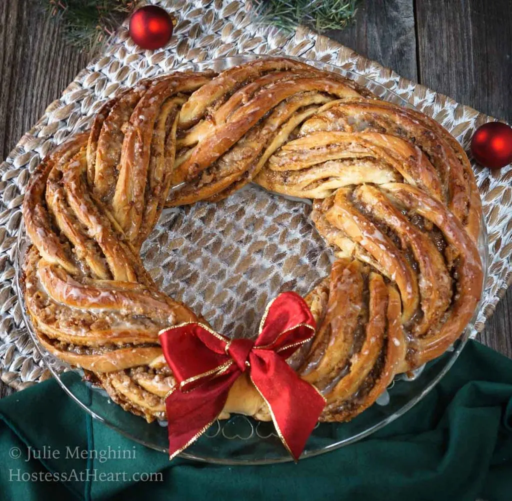 Sweet Nut Holiday Bread baked in the shape of a wreath and garnished with a red bow. A green napkin and a weaved placemat sit under the wreath. Two Christmas bulbs sit in the background.