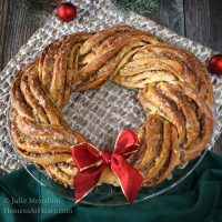 Sweet Nut Holiday Bread baked in the shape of a wreath and garnished with a red bow. A green napkin and a weaved placemat sit under the wreath. Two Christmas bulbs sit in the background.