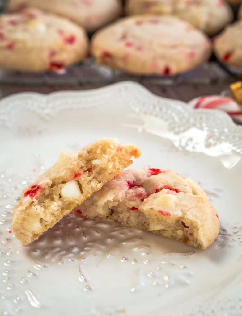 a cookie split in half showing white chocolate chips and pieces of peppermint on a white plate with a rack of cookies behind it.