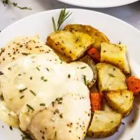 A chicken breast with a lemon herb sauce sitting on a white plate next to roasted potatoes. The plate sits over a green napkin with herbs in the background.
