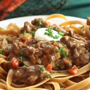 Close up side view of a white plate of beef stroganoff on a white plate with a spoonful of sour cream sitting on top of it.