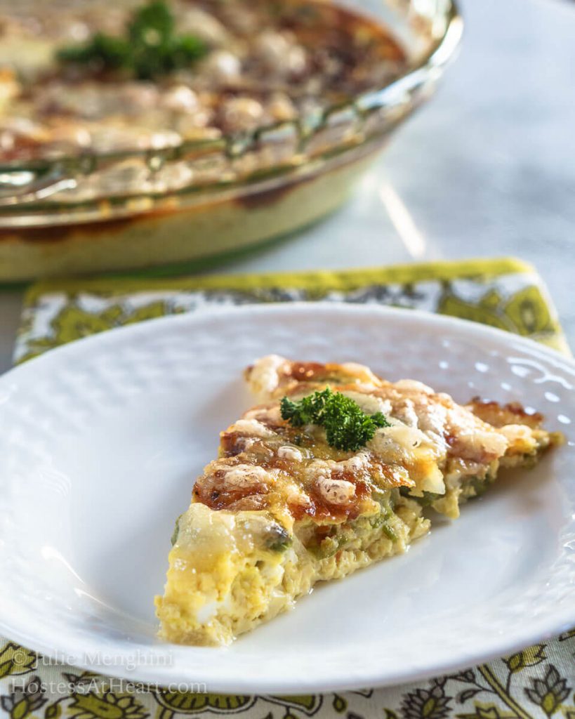 A slice of Crustless 3 Cheese Asparagus Quiche sitting on a white plate in front of the pie plate of quiche.