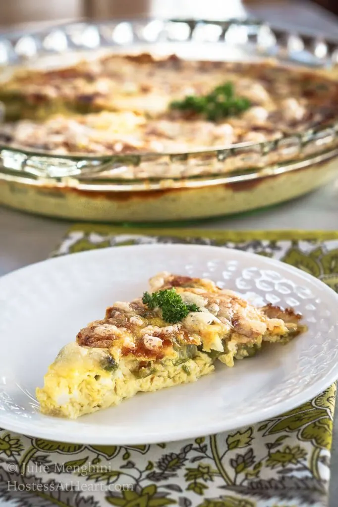 A slice of Crustless 3 Cheese Asparagus Quiche sitting on a white plate in front of the pie plate of quiche.